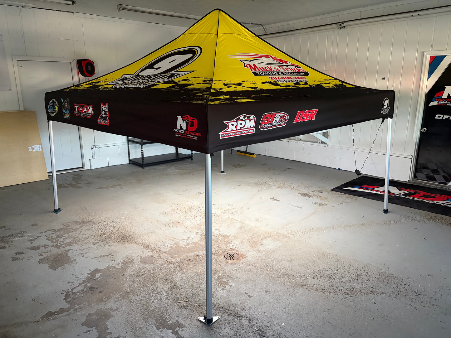 A pop-up canopy tent with a yellow and black racing design, featuring sponsor logos such as RPM, GO, and NDR, set up in a garage.