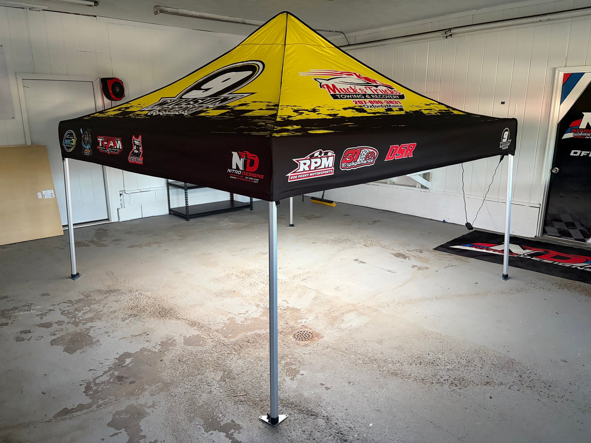 A pop-up canopy tent with a yellow and black racing design, featuring sponsor logos such as RPM, GO, and NDR, set up in a garage.